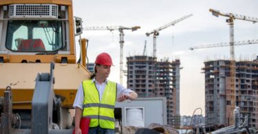 A construction manager stands on a jobsite and looks at his watch. Cranes and heavy equipment are in the background.