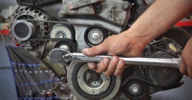 A mechanic's hands use a torque wrench on a vehicle's engine. Various components are visible, including one of its belts.