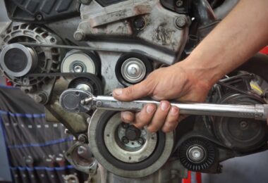 A mechanic's hands use a torque wrench on a vehicle's engine. Various components are visible, including one of its belts.