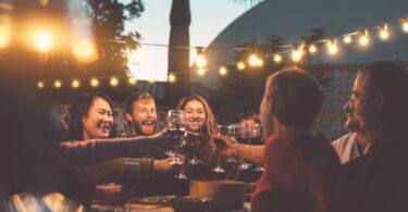 A group of happy people toasting with glasses of red wine over an outdoor table while at a sunset dinner party.