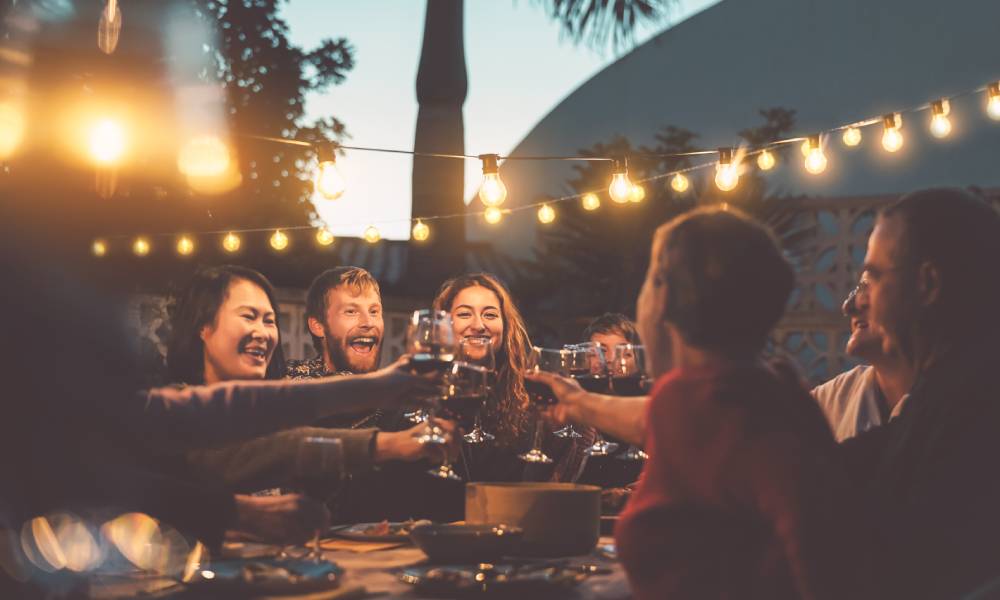 A group of happy people toasting with glasses of red wine over an outdoor table while at a sunset dinner party.