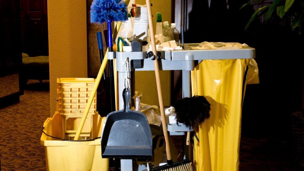 A collection of essential cleaning supplies for businesses displayed in a dimly lit room, including mops, buckets, brooms, and various cleaning agents on a janitorial cart. 