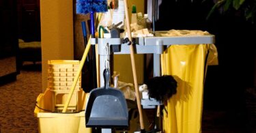 A collection of essential cleaning supplies for businesses displayed in a dimly lit room, including mops, buckets, brooms, and various cleaning agents on a janitorial cart.