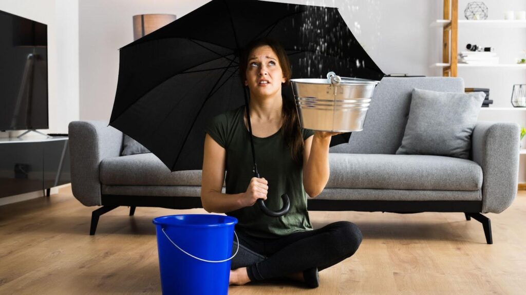 Woman sitting on the floor of her living room holding a bucket to catch water from a leak in the ceiling, using an umbrella to shield herself, illustrating the need for proactive measures to protect homes from water damage.