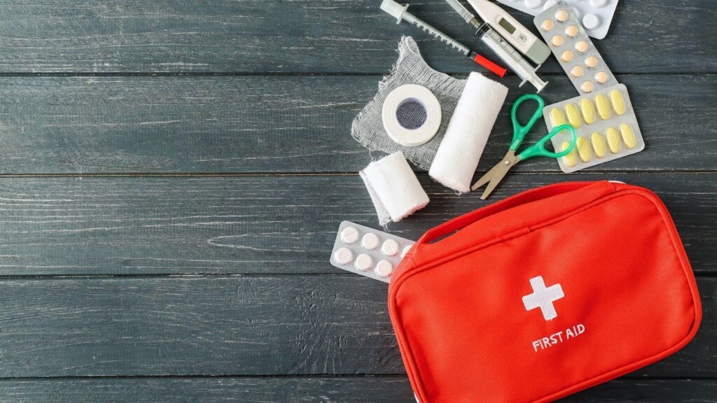 Essential first aid supplies including bandages, scissors, tape, and medications displayed alongside a red first aid kit on a wooden background 