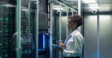 A woman wearing a lanyard at a data center server rack inspecting the hardware while holding a tablet.