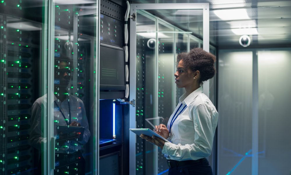 A woman wearing a lanyard at a data center server rack inspecting the hardware while holding a tablet.