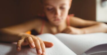 A young girl in a tank top using her right hand to read braille from an open book filled with white pages.