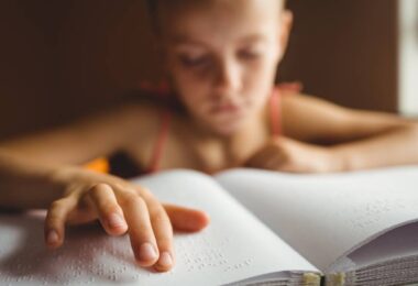 A young girl in a tank top using her right hand to read braille from an open book filled with white pages.