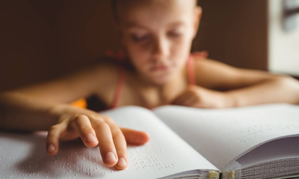 A young girl in a tank top using her right hand to read braille from an open book filled with white pages.