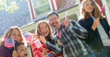 A family of five standing outside a red house on a sunny day. They are all smiling and waving small American flags.