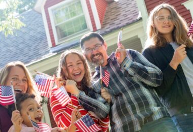 A family of five standing outside a red house on a sunny day. They are all smiling and waving small American flags.