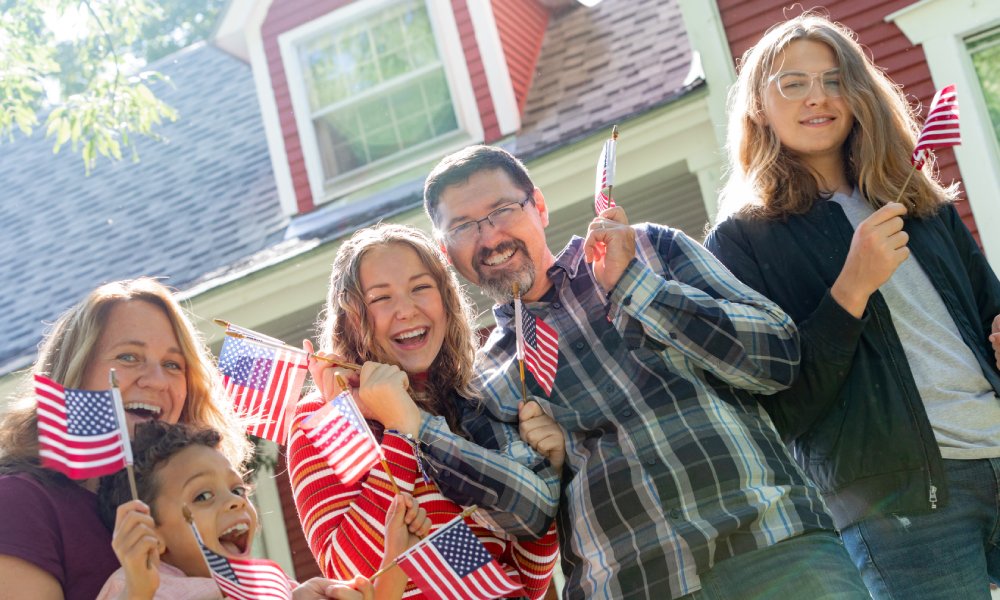 A family of five standing outside a red house on a sunny day. They are all smiling and waving small American flags.