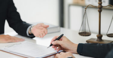 Two people wearing suits, sitting at a desk in front of a set of scales representing the law. One is signing paperwork.