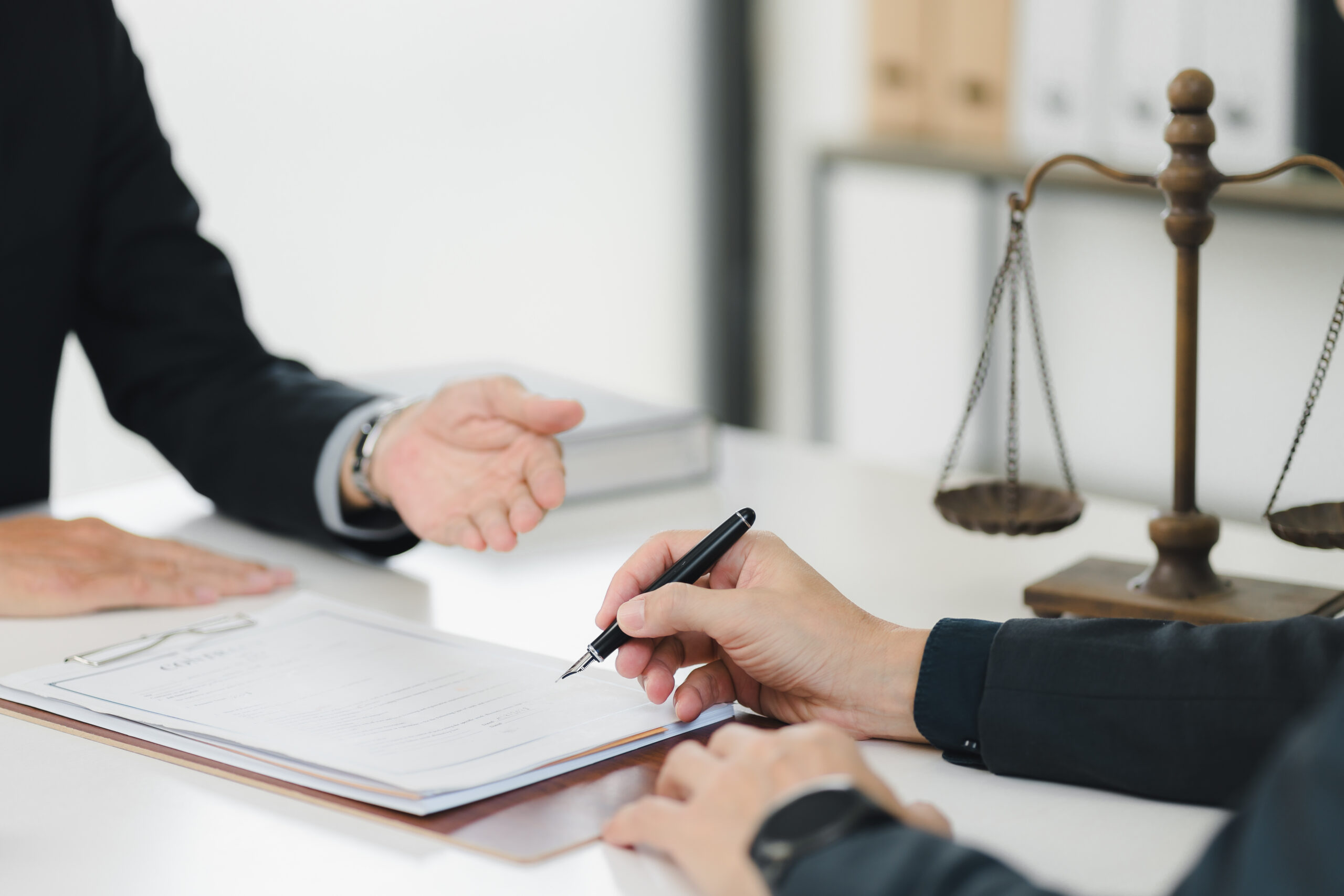 Two people wearing suits, sitting at a desk in front of a set of scales representing the law. One is signing paperwork.