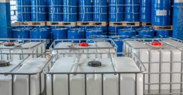 A group of white plastic storage tanks inside of a building. There are blue barrels behind the storage tanks.