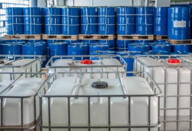 A group of white plastic storage tanks inside of a building. There are blue barrels behind the storage tanks.