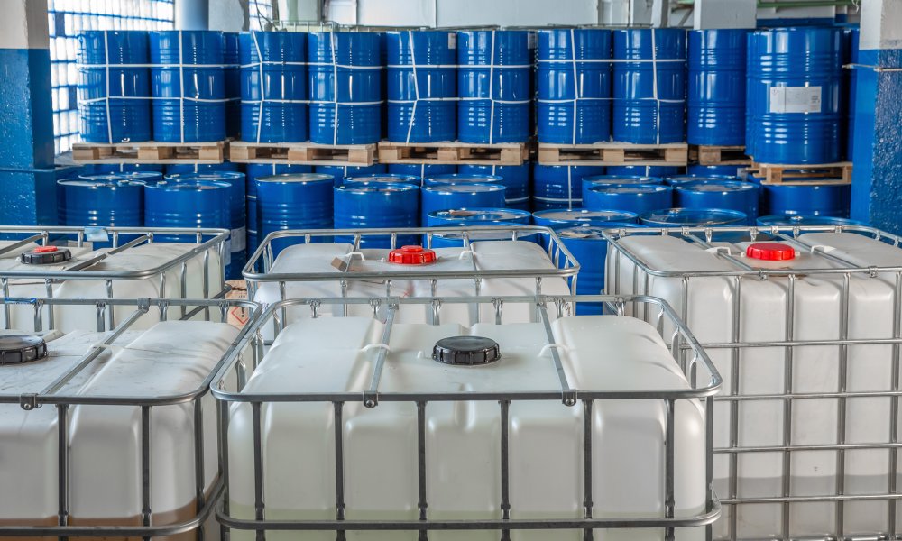 A group of white plastic storage tanks inside of a building. There are blue barrels behind the storage tanks.