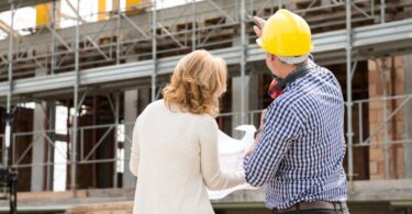 Two people standing in front of a building under construction. One of them is wearing a hard hat, pointing at the structure.