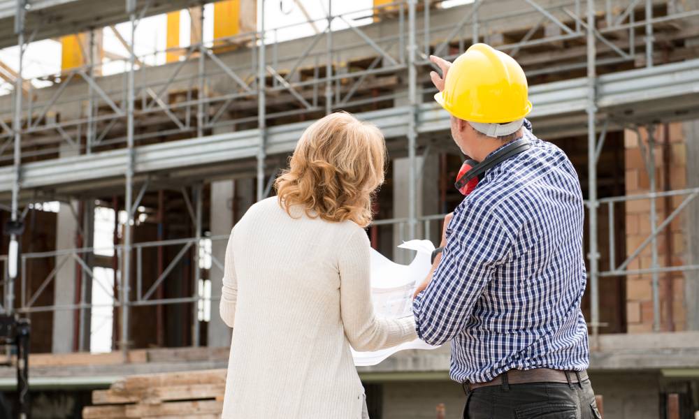 Two people standing in front of a building under construction. One of them is wearing a hard hat, pointing at the structure.