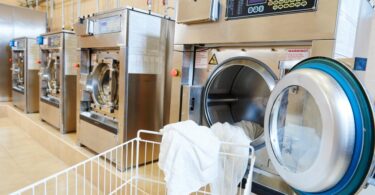 A cart sits in front of a commercial laundry machine with the door open and a white sheet hanging out of the door.