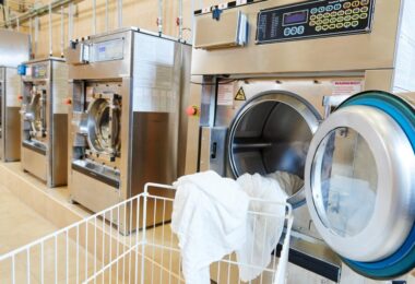 A cart sits in front of a commercial laundry machine with the door open and a white sheet hanging out of the door.