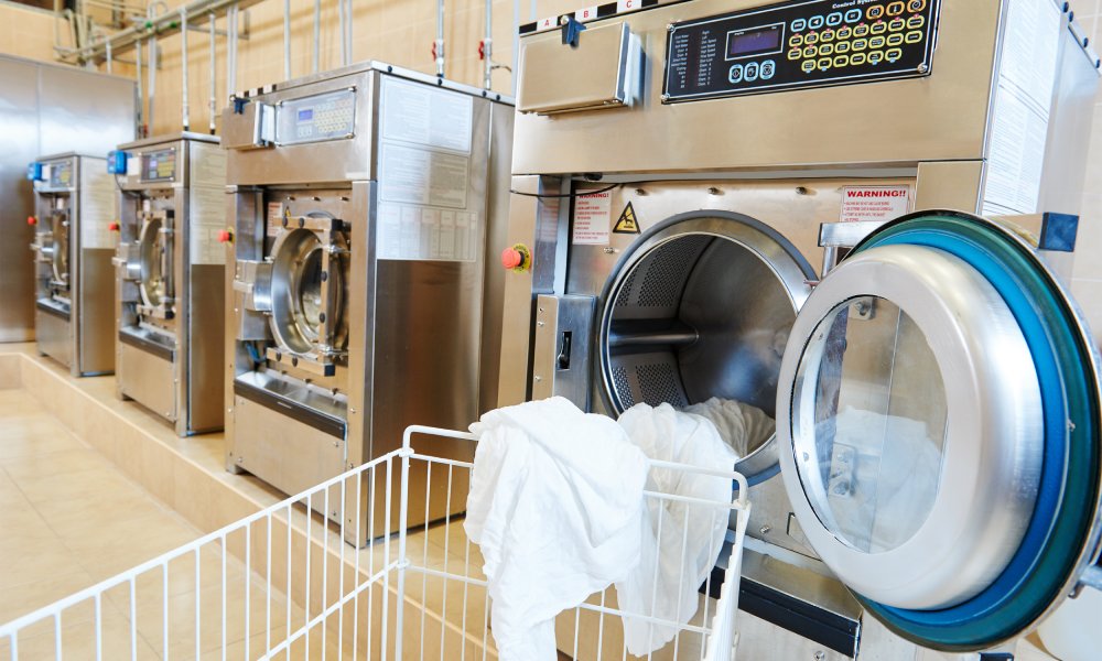 A cart sits in front of a commercial laundry machine with the door open and a white sheet hanging out of the door.