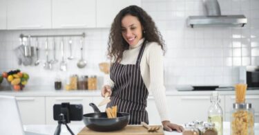 A young woman is wearing a striped apron and cooking in a kitchen. Her laptop and smartphone are on the counter.