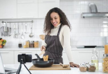 A young woman is wearing a striped apron and cooking in a kitchen. Her laptop and smartphone are on the counter.