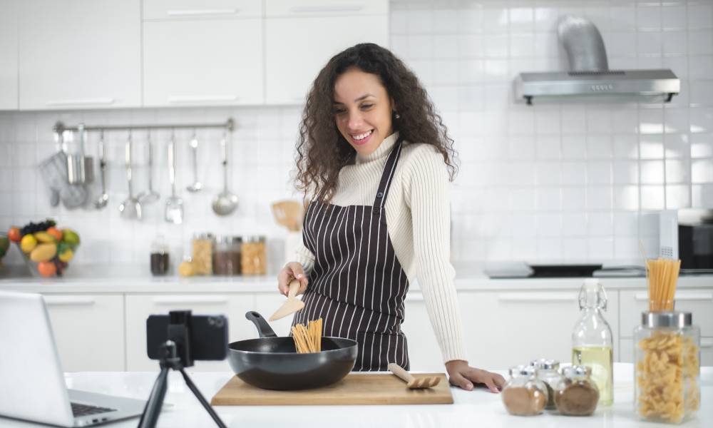 A young woman is wearing a striped apron and cooking in a kitchen. Her laptop and smartphone are on the counter.