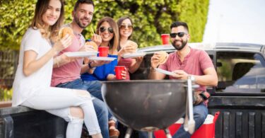 A group of young adult friends sitting and eating in the bed of a truck with a charcoal grill in the foreground.