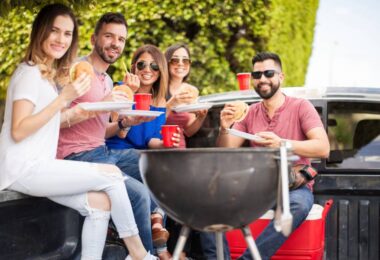 A group of young adult friends sitting and eating in the bed of a truck with a charcoal grill in the foreground.