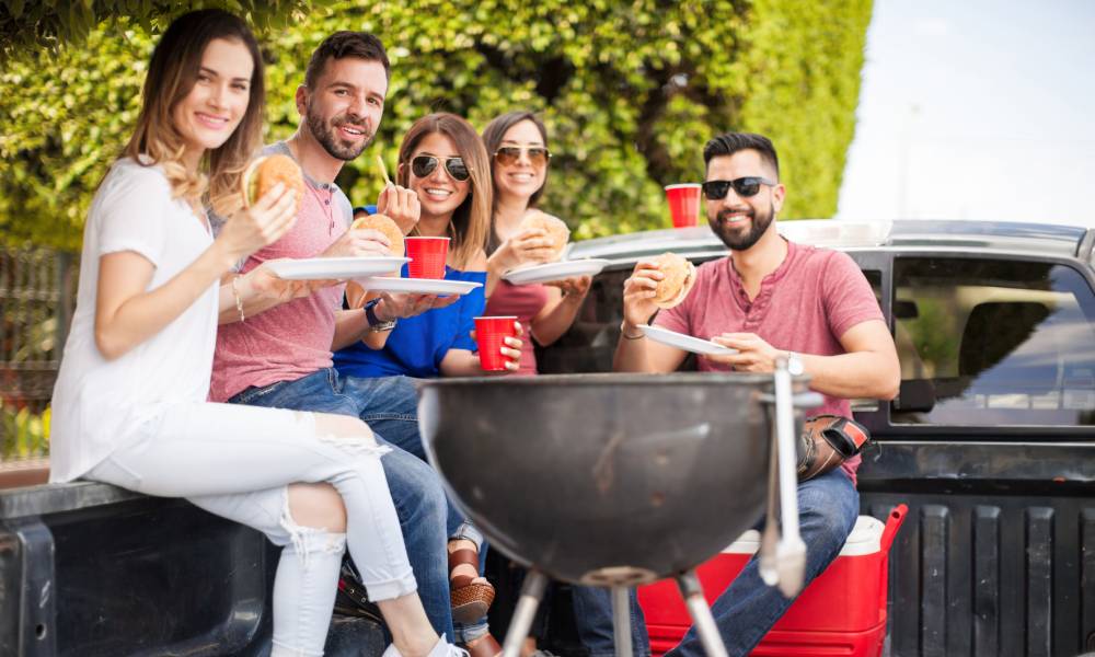 A group of young adult friends sitting and eating in the bed of a truck with a charcoal grill in the foreground.