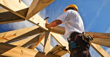 A man in a white t-shirt, yellow hard hat, and cargo pants hammers a wooden structure on a sunny day.