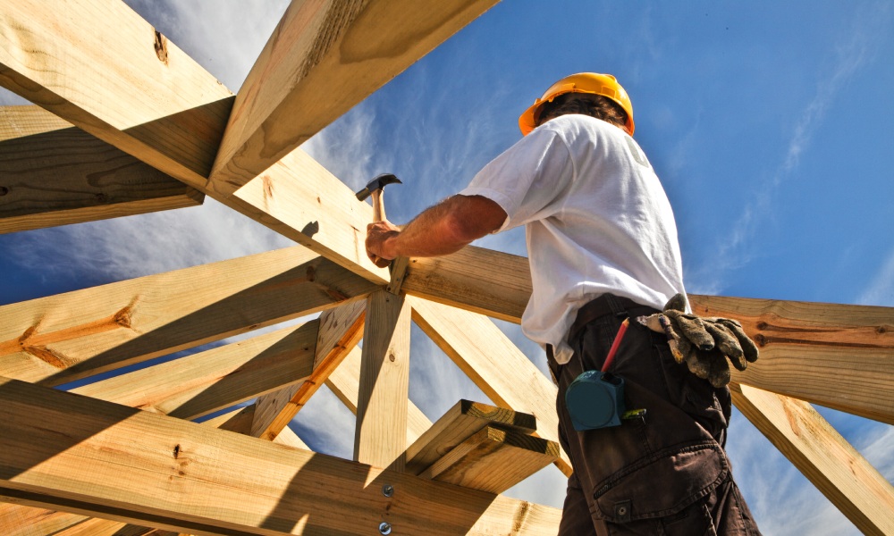 A man in a white t-shirt, yellow hard hat, and cargo pants hammers a wooden structure on a sunny day.