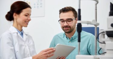 A smiling eye doctor holds a tablet device while explaining something to her male patient in an exam room.