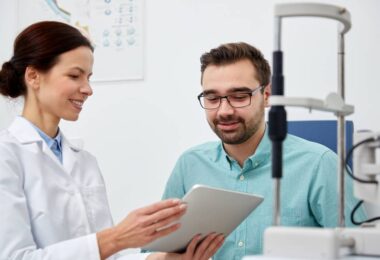 A smiling eye doctor holds a tablet device while explaining something to her male patient in an exam room.