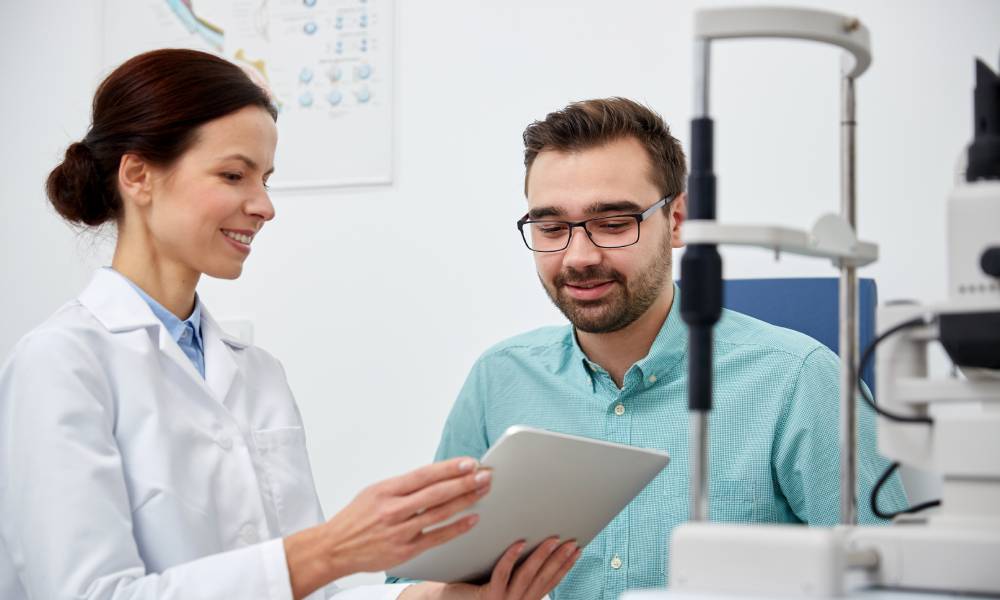 A smiling eye doctor holds a tablet device while explaining something to her male patient in an exam room.