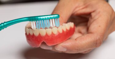 An individual is holding a green toothbrush with blue and white bristles and brushing a pair of dentures.