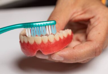 An individual is holding a green toothbrush with blue and white bristles and brushing a pair of dentures.