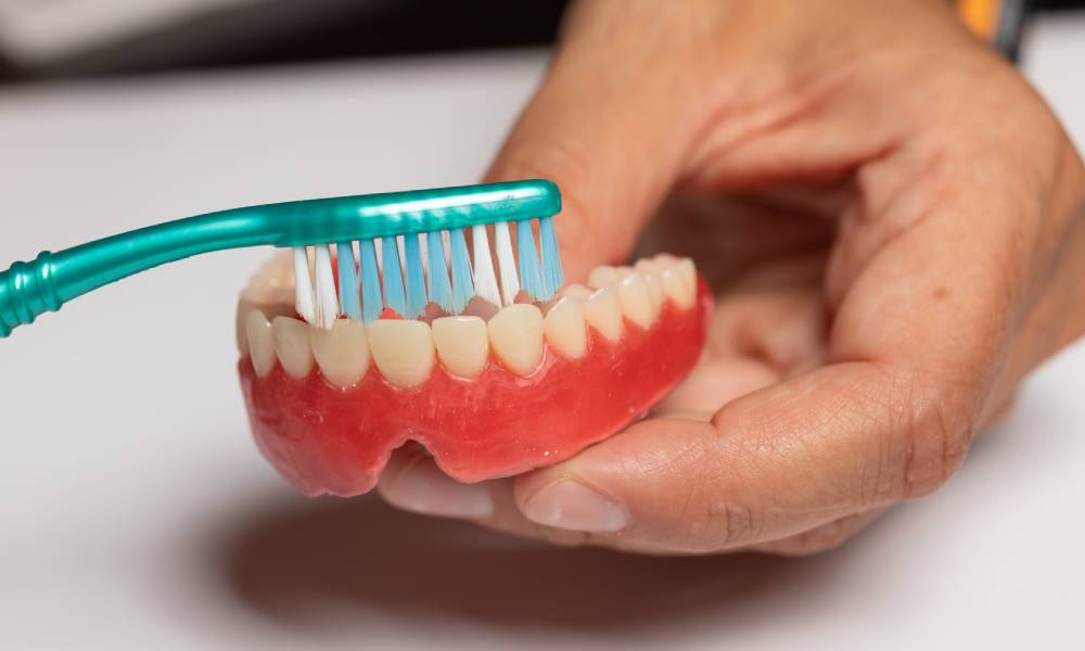 An individual is holding a green toothbrush with blue and white bristles and brushing a pair of dentures.