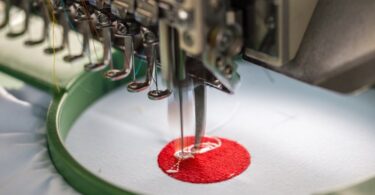 A close-up of an embroidery machine's needle in use. The machine is making a red circle with white in the middle.