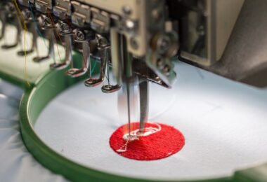 A close-up of an embroidery machine's needle in use. The machine is making a red circle with white in the middle.