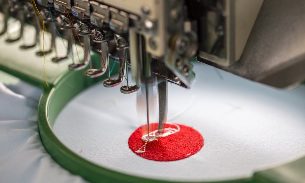 A close-up of an embroidery machine's needle in use. The machine is making a red circle with white in the middle.