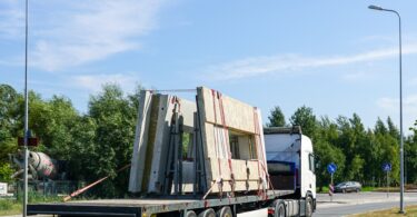 Precast concrete walls strapped to the bed of a long flatbed truck that is driving down a road.