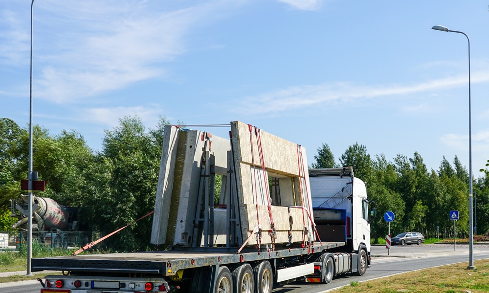 Precast concrete walls strapped to the bed of a long flatbed truck that is driving down a road.