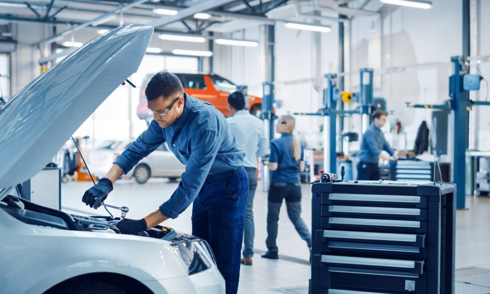 A close-up of a male car mechanic wearing safety gear and working on a silver vehicle in a commercial garage.