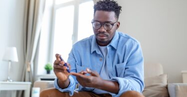 A man wearing a blue shirt, khaki pants, and glasses is using a blue tool to prick his fingertip while sitting on a couch.