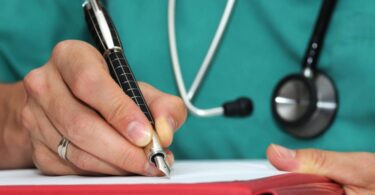 Close-up on the hands of a woman writing with a black pen on a red folder and a sheet of paper, wearing a stethoscope and scrubs.