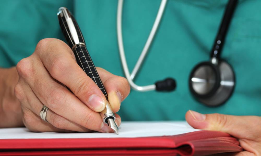 Close-up on the hands of a woman writing with a black pen on a red folder and a sheet of paper, wearing a stethoscope and scrubs.